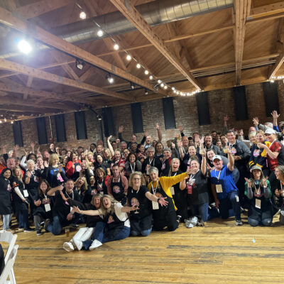 A large group of people posing together in a celebratory manner at an indoor chili cook-off team building. The participants are smiling, waving, and cheering while wearing aprons with chili cook-off branding. thumbnail
