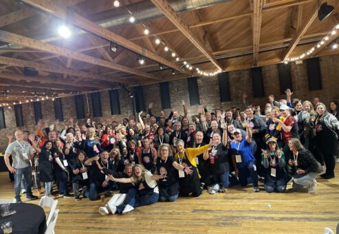 A large group of people posing together in a celebratory manner at an indoor chili cook-off team building. The participants are smiling, waving, and cheering while wearing aprons with chili cook-off branding.