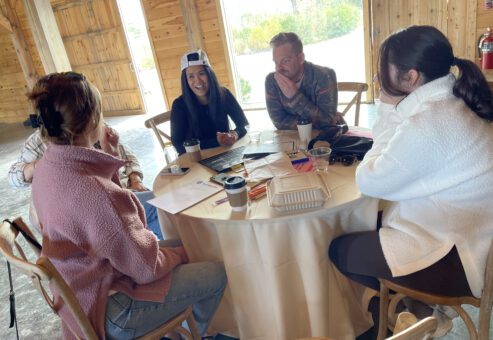 Group of four people sitting around a round table engaged in a discussion, with papers, coffee cups, and a laptop, capturing a collaborative team building moment.