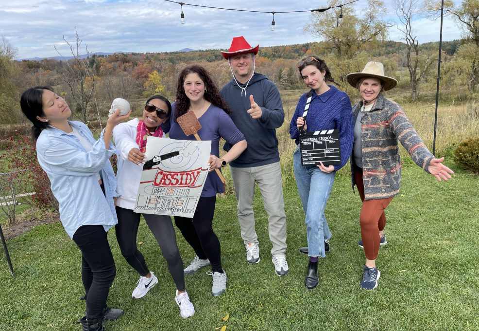A group of six people pose outdoors, holding various props like a film clapperboard, a poster, and a red cowboy hat, participating in a creative team building activity themed around film production and storytelling.