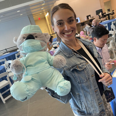 A smiling participant holds a teddy bear dressed as a doctor in light blue scrubs and a matching face mask. The bear is part of the Team Teddy Rescue Bear team building event, where participants create and dress up teddy bears for charity. thumbnail