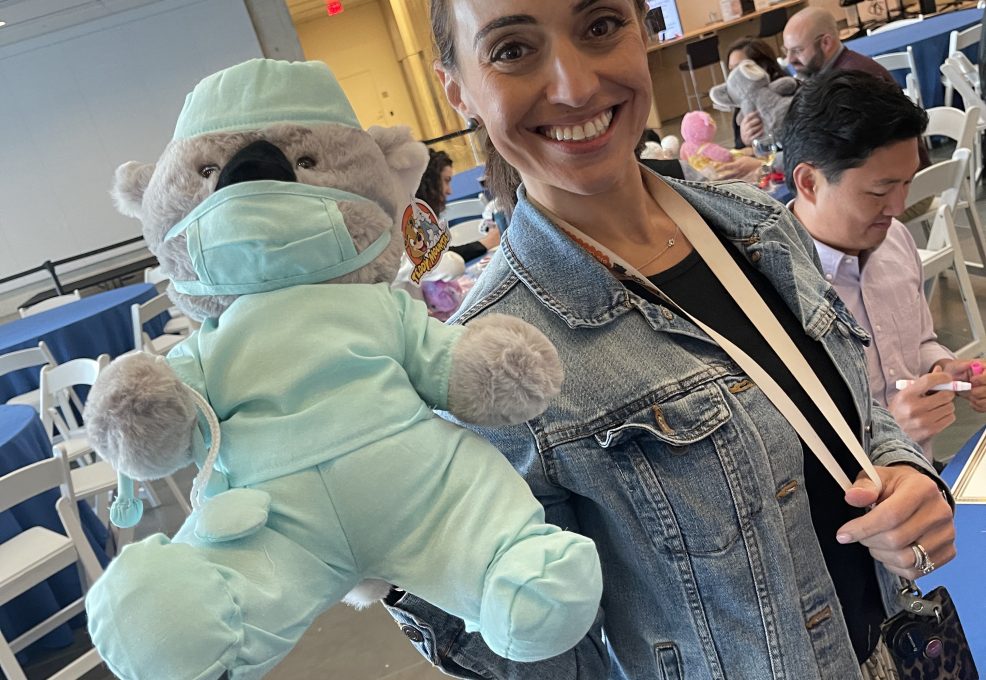A smiling participant holds a teddy bear dressed as a doctor in light blue scrubs and a matching face mask. The bear is part of the Team Teddy Rescue Bear team building event, where participants create and dress up teddy bears for charity.