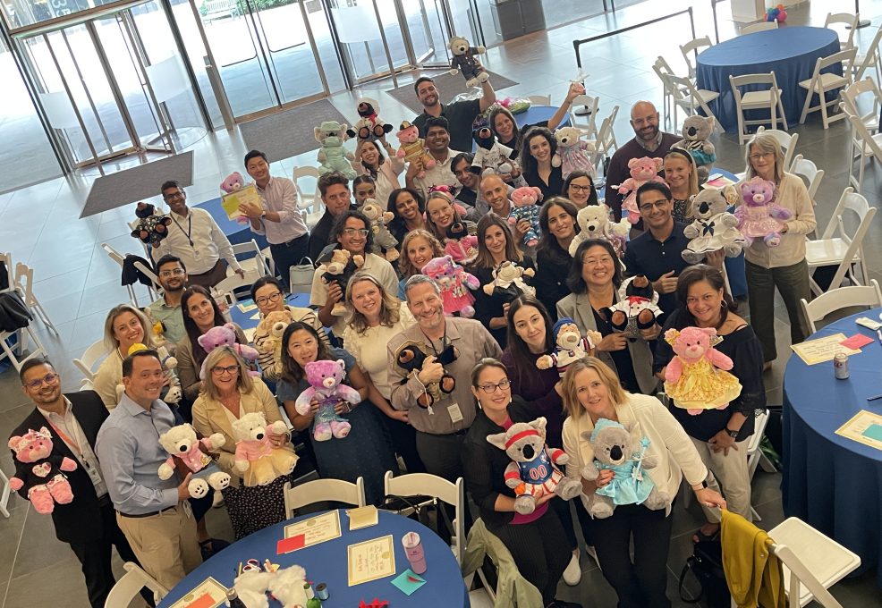 A large group of participants proudly holding their completed teddy bears at the end of the Team Teddy Rescue Bear event. The participants, arranged in a group around circular tables, smile and showcase the colorful, dressed-up teddy bears they made as part of the team building CSR event.