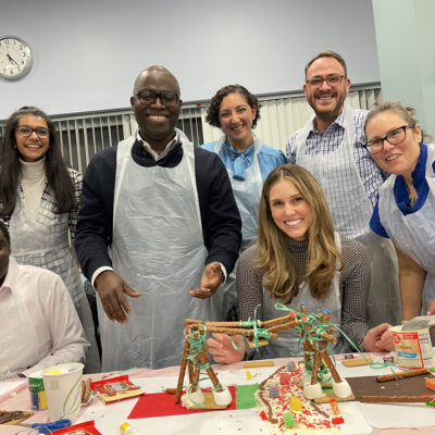A smiling team proudly stands behind their creative chocolate bridge structure during The Chocolate Challenge team building event. Each team member is wearing aprons, and the table is filled with candy decorations and construction materials, showing their collaborative efforts. thumbnail