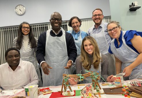 A smiling team proudly stands behind their creative chocolate bridge structure during The Chocolate Challenge team building event. Each team member is wearing aprons, and the table is filled with candy decorations and construction materials, showing their collaborative efforts.