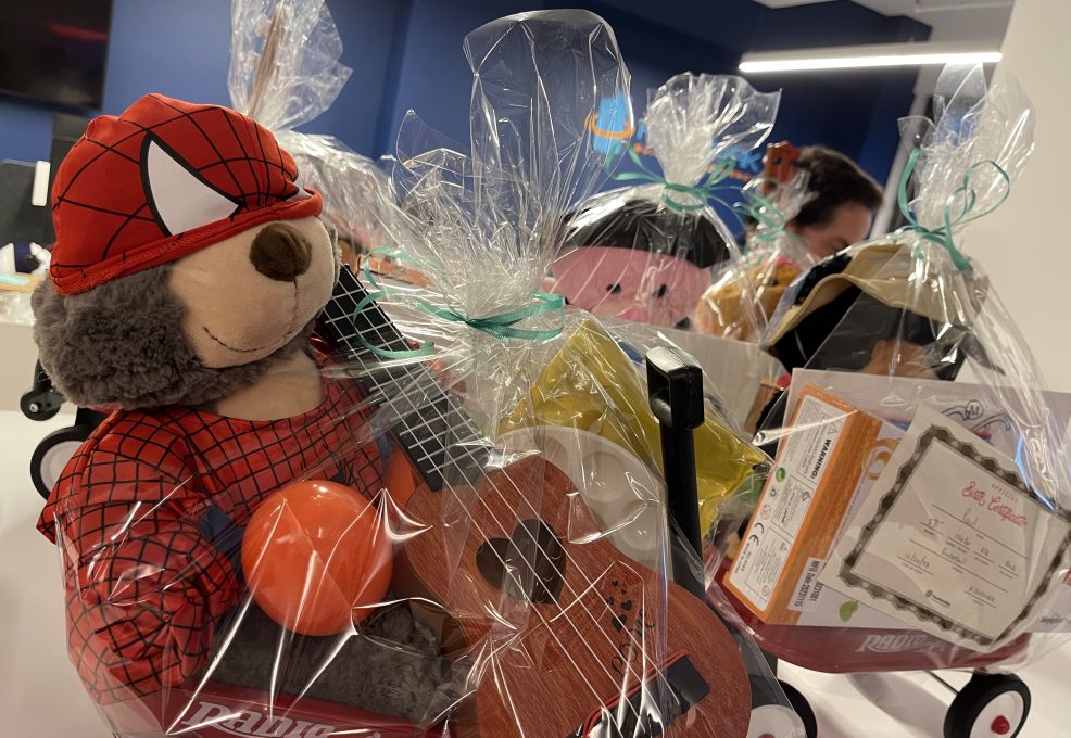 A close-up image of a red Radio Flyer wagon filled with a gift set for charity. The wagon includes a stuffed teddy bear dressed in a superhero outfit, a small ukulele, a maraca, and various art supplies. The entire wagon is wrapped in clear gift wrap, ready for donation at the Toys for Tykes ‘Arts & Music’ team building event.
