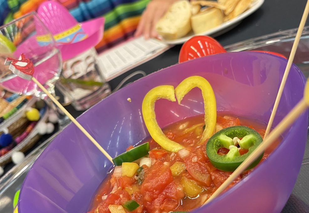 A colorful bowl of salsa garnished with heart-shaped yellow peppers and jalapeños at a Salsa Showdown team building event, with participants wearing vibrant striped outfits.