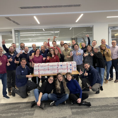 Team poses with completed care packages during the Operation Military Care team building event, ready to be sent to military personnel overseas. thumbnail