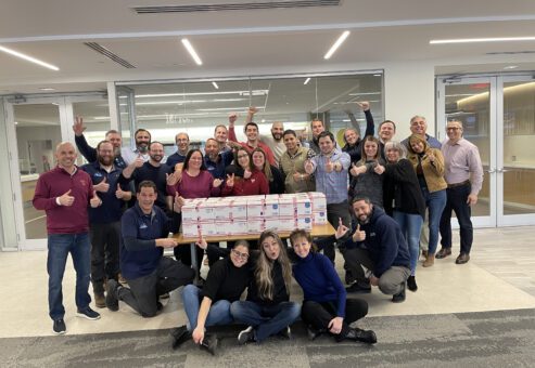 Team poses with completed care packages during the Operation Military Care team building event, ready to be sent to military personnel overseas.