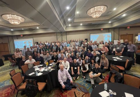 A large group gathers together after successfully completing an Operation Military Care team building event, having packed care packages for U.S. troops.