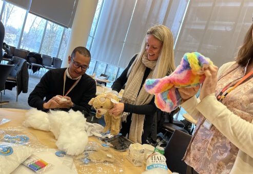 Three participants in the Team Teddy Rescue Bear team building activity are gathered around a table filled with teddy bear stuffing and various craft supplies. A woman is holding a colorful rainbow-patterned teddy bear, while a man next to her is assembling another bear.