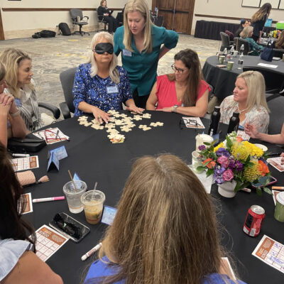 A group of women participating in a team building activity sitting around a round table. One woman is wearing a blindfold and feeling puzzle pieces in front of her. Other participants are observing and giving instructions to hep her complete the challenge. thumbnail