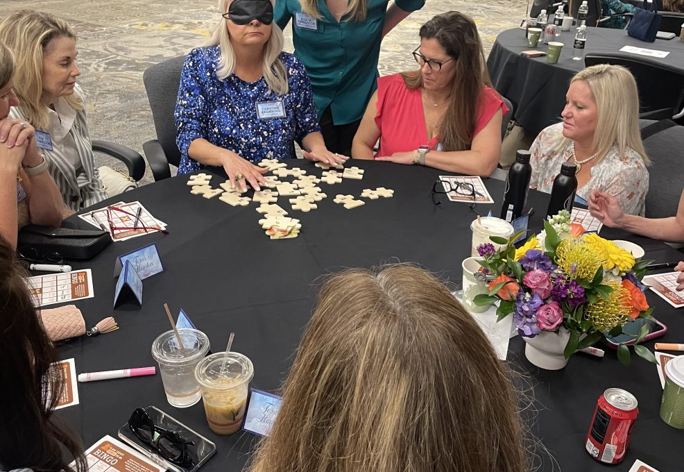 A group of women participating in a team building activity sitting around a round table. One woman is wearing a blindfold and feeling puzzle pieces in front of her. Other participants are observing and giving instructions to hep her complete the challenge.