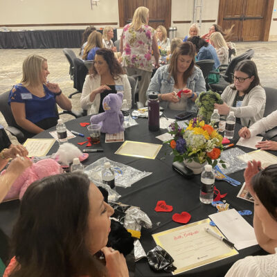 A group of women seated at a round table, engaged in a creative activity. Some are working on decorating or assembling colorful teddy bears, while others are chatting or focused on materials in front of them. The table is adorned with a vase of flowers, water bottles, and various teddy bear-making supplies, such as fabric hearts and stuffing. The room in the background shows more tables with participants working together in CSR team building program. thumbnail