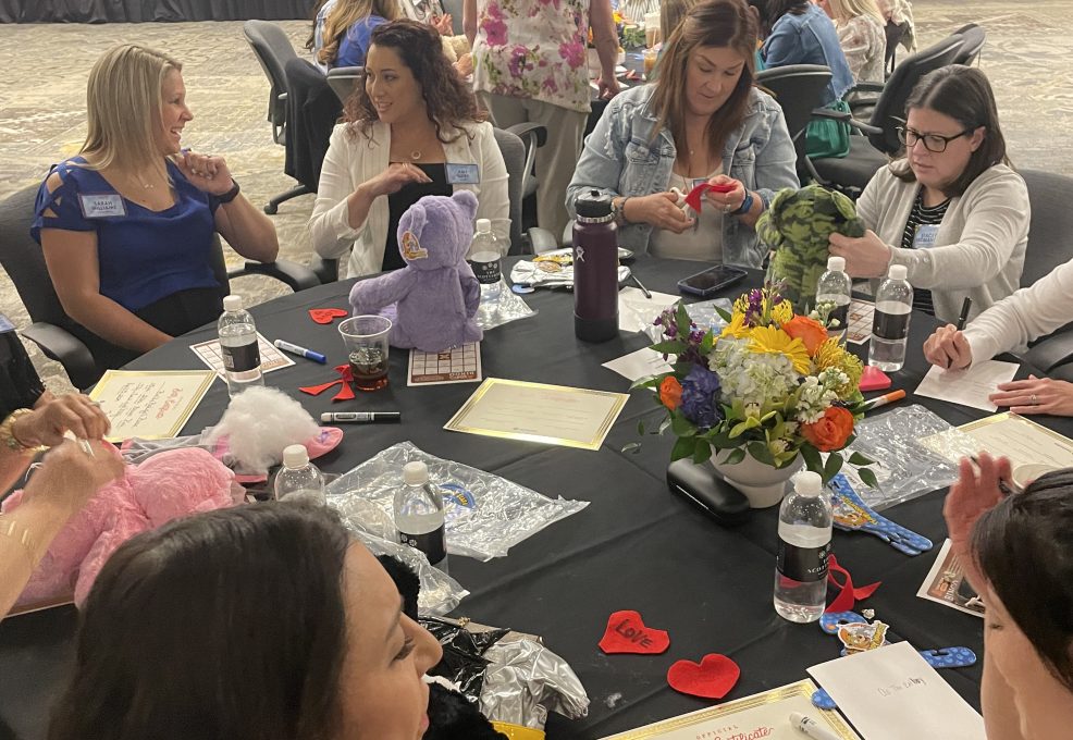 A group of women seated at a round table, engaged in a creative activity. Some are working on decorating or assembling colorful teddy bears, while others are chatting or focused on materials in front of them. The table is adorned with a vase of flowers, water bottles, and various teddy bear-making supplies, such as fabric hearts and stuffing. The room in the background shows more tables with participants working together in CSR team building program.
