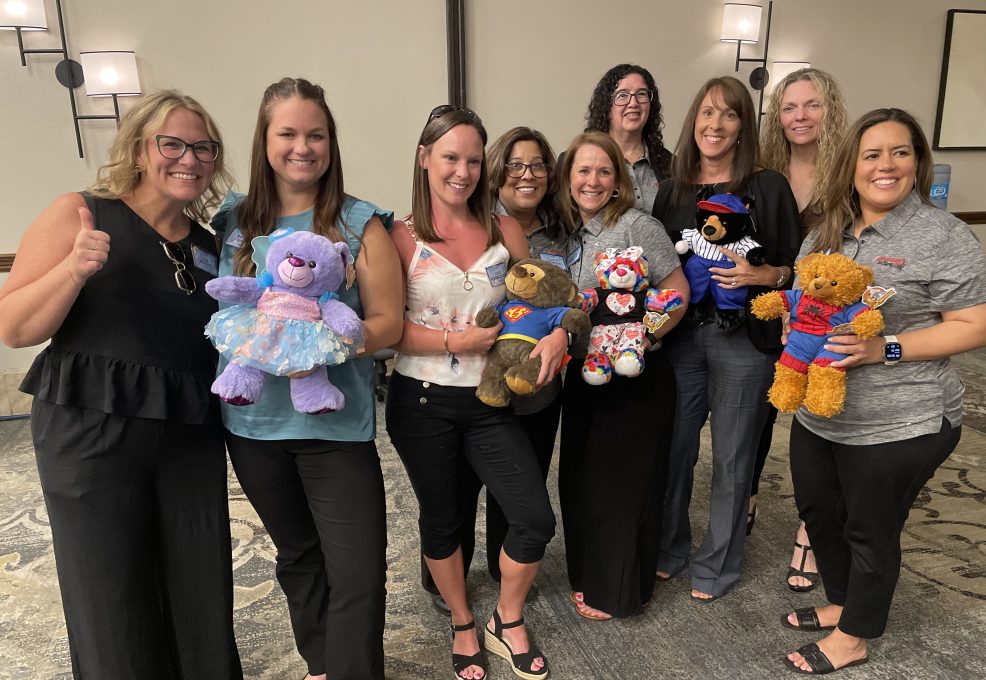 A group of eight smiling women posing together, holding customized teddy bears. Each teddy bear is dressed uniquely, with some wearing superhero costumes, a tutu, or colorful accessories. The women are standing in a conference room, showing off their creations proudly as part of a team building activity.