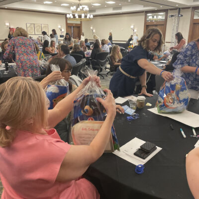 A group of women at a table are carefully assembling gift bags during a large event. The bags contain stuffed animals, certificates, and other items, for a charitable team building program. The room is filled with other participants engaged in similar activities. thumbnail