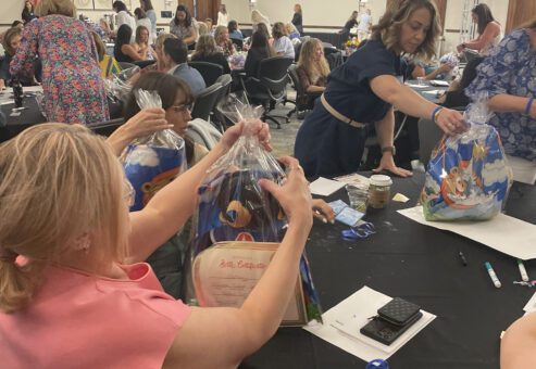 A group of women at a table are carefully assembling gift bags during a large event. The bags contain stuffed animals, certificates, and other items, for a charitable team building program. The room is filled with other participants engaged in similar activities.