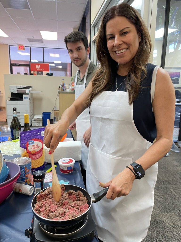 A woman and a man participate in a culinary team building event. The woman is smiling as she stirs a pan filled with ingredients while wearing a white apron. Various food items like cheese and sour cream are visible on the table in front of them. The man behind her is also wearing an apron, standing by the food preparation area.