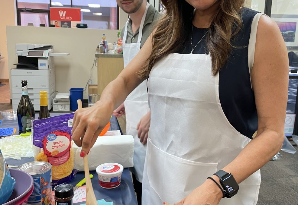 A woman and a man participate in a culinary team building event. The woman is smiling as she stirs a pan filled with ingredients while wearing a white apron. Various food items like cheese and sour cream are visible on the table in front of them. The man behind her is also wearing an apron, standing by the food preparation area.