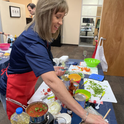A woman is participating in the team building chili cook-off. She's wearing a red apron is smiling while cooking at a table filled with ingredients and kitchen tools. She is holding a small container to add seasoning to a pan filled with chopped vegetables and sausages. On the table, there are various ingredients, including cans, chopped onions, cilantro, a bottle of cooking oil, and a glass of red wine. thumbnail