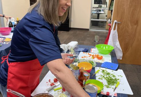 A woman is participating in the team building chili cook-off. She's wearing a red apron is smiling while cooking at a table filled with ingredients and kitchen tools. She is holding a small container to add seasoning to a pan filled with chopped vegetables and sausages. On the table, there are various ingredients, including cans, chopped onions, cilantro, a bottle of cooking oil, and a glass of red wine.