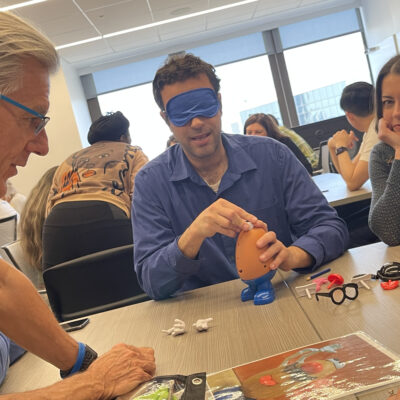 A group of people participates in a Toys for Tykes team building activity. One man is blindfolded while assembling a Mr. Potato Head toy, with other team members observing and giving instructions, creating a fun and collaborative atmosphere. thumbnail
