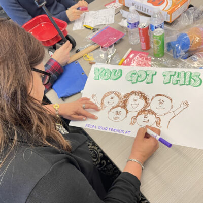 A person seated at a table working on a hand-drawn sign that reads 'You Got This!' The drawing includes smiling faces of people and the words 'From your friends at...' as part of the message. Surrounding the person are various arts and crafts supplies, this is part of a team building event. thumbnail