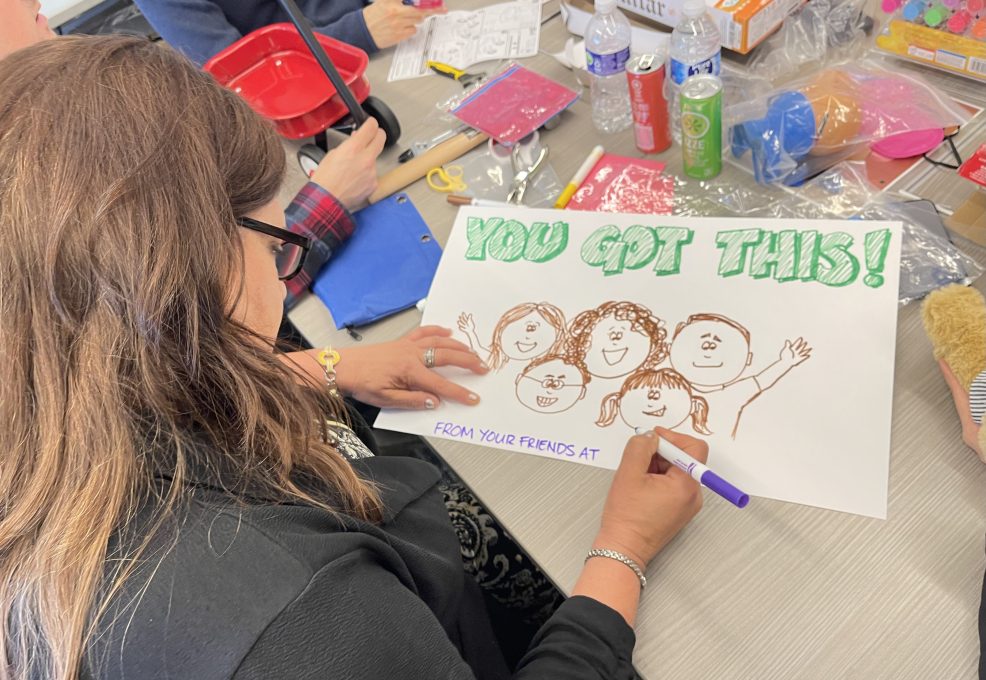 A person seated at a table working on a hand-drawn sign that reads 'You Got This!' The drawing includes smiling faces of people and the words 'From your friends at...' as part of the message. Surrounding the person are various arts and crafts supplies, this is part of a team building event.