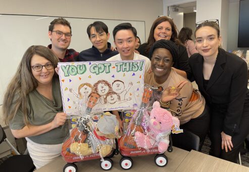 A team of seven people stands around a table holding two small red wagons decorated with teddy bears and musical instruments, all intended for donation. One team member holds a sign reading 'You Got This!' with a fun drawing of people cheering. The group is smiling and posing for the camera, clearly proud of their work at the Toys for Tykes ‘Arts & Music’ team building event.