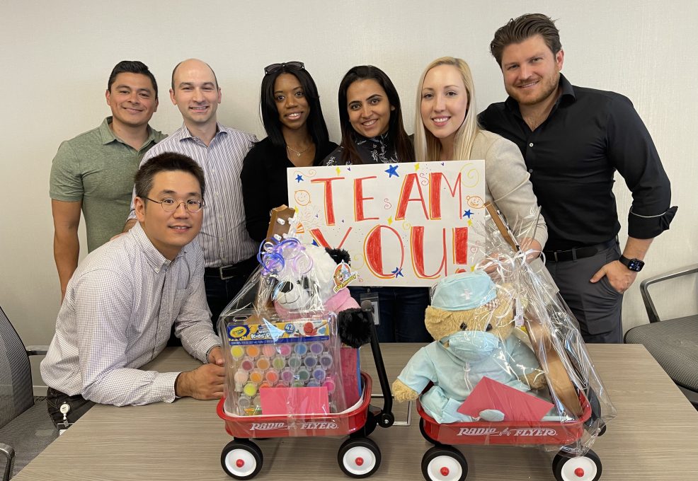 A team of six people proudly showcases their completed toy wagons filled with stuffed animals, arts and crafts supplies, and a sign that reads 'TEAM YOU!' as part of a Toys for Tykes team building event.