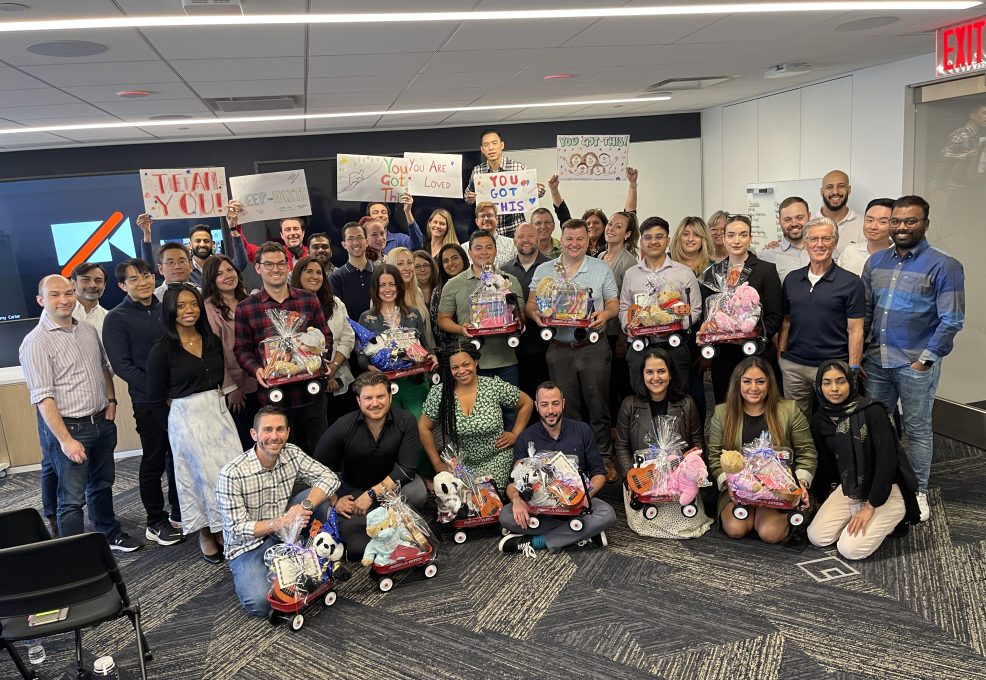 A large group of people smiling and posing together in a modern office space. They are holding and sitting with small red Radio Flyer wagons filled with various toys, teddy bears, and musical instruments, all wrapped in gift wrap. Behind them, some participants are holding up hand-drawn signs with motivational messages like 'You Got This!' This photo captures the conclusion of the Toys for Tykes ‘Arts & Music’ CSR team building event.