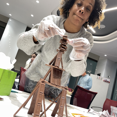 A participant in the Chocolate Challenge team building event carefully stacks chocolate bars to create a stable structure, focusing on precision and creativity. thumbnail