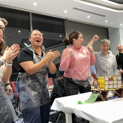 Team members celebrate their success in the Chocolate Challenge team building event, excitedly clapping after constructing their chocolate bridge. thumbnail