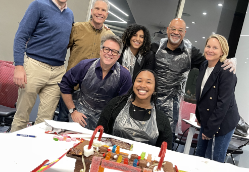 A team proudly poses with their creative, candy-decorated chocolate bridge after completing the Chocolate Challenge team building event, showcasing teamwork and fun.
