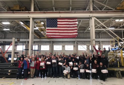 Large group of participants proudly holding care packages for military personnel, standing beneath the American flag during the Operation Military Care team building event.