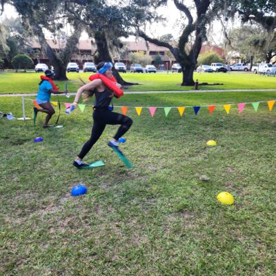 A participant wearing a life vest and flippers runs through an obstacle course during the Outrageous Games team building event, showcasing fun and friendly competition. thumbnail