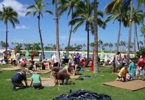 Teams constructing cardboard boats on a sunny lawn surrounded by palm trees during the Build-A-Boat team building event.