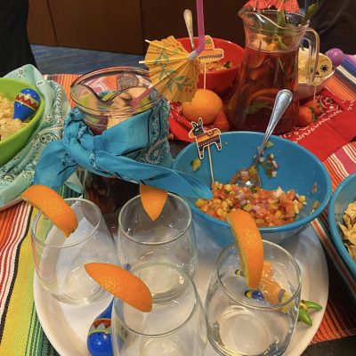 A festive spread featuring glasses garnished with orange slices, a pitcher of colorful drink, fresh salsa, and bowls of chips, all part of the Salsa Showdown team building event. thumbnail