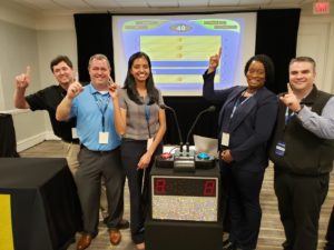 A team of five participants posing with enthusiasm and pointing to the screen, showing a game show setup during the Corporate Feud team building activity, highlighting team spirit, competition, and fun.
