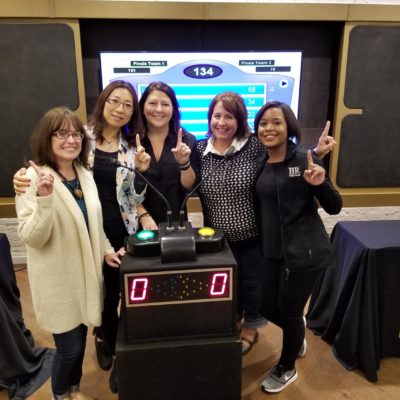 Five participants smiling and holding up their index fingers in front of a game show podium, celebrating their involvement in the Corporate Feud team building activity, with a game board displayed in the background. thumbnail