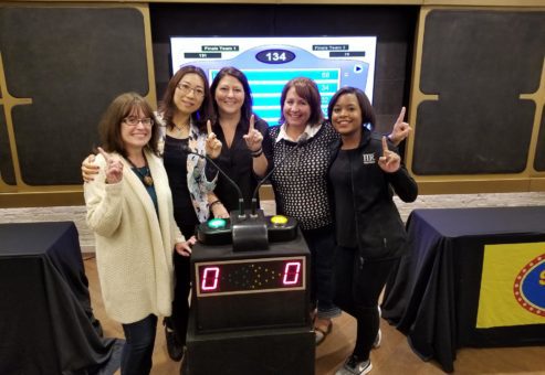 Five participants smiling and holding up their index fingers in front of a game show podium, celebrating their involvement in the Corporate Feud team building activity, with a game board displayed in the background.