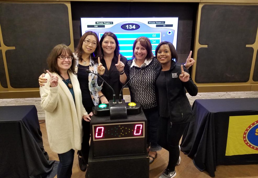 Five participants smiling and holding up their index fingers in front of a game show podium, celebrating their involvement in the Corporate Feud team building activity, with a game board displayed in the background.