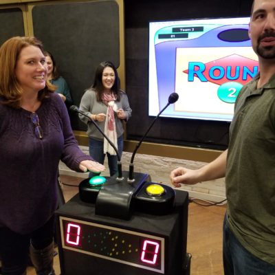 Two participants standing at a game show podium with buzzers, smiling and preparing to answer during the survey game show team building event, with other participants watching in the background. thumbnail