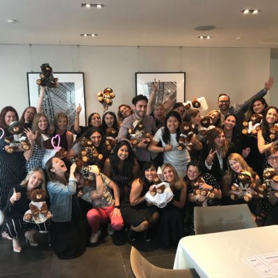 A large group of enthusiastic participants, smiling and posing with teddy bears they have assembled during a team building event. The group, made up of men and women, holds up their brown teddy bears. Many are raising their hands or posing playfully, creating a joyful and celebratory atmosphere. thumbnail