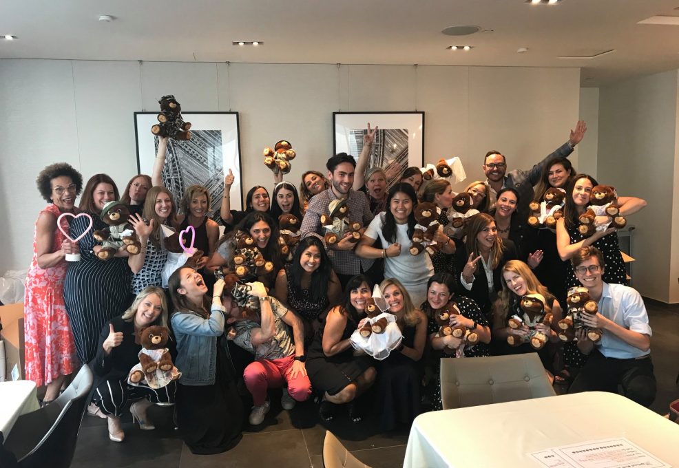 A large group of enthusiastic participants, smiling and posing with teddy bears they have assembled during a team building event. The group, made up of men and women, holds up their brown teddy bears. Many are raising their hands or posing playfully, creating a joyful and celebratory atmosphere.