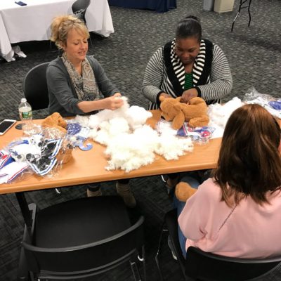 Three women seated at a table during a teddy bear assembly team building event. They are focused on stuffing and creating teddy bears from kits that include plush materials, stuffing, and small accessories. Various bear parts and materials are scattered on the table, and they appear to be enjoying the hands-on activity. thumbnail