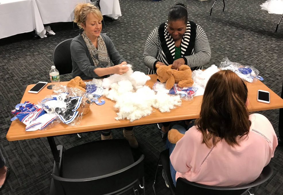 Three women seated at a table during a teddy bear assembly team building event. They are focused on stuffing and creating teddy bears from kits that include plush materials, stuffing, and small accessories. Various bear parts and materials are scattered on the table, and they appear to be enjoying the hands-on activity.