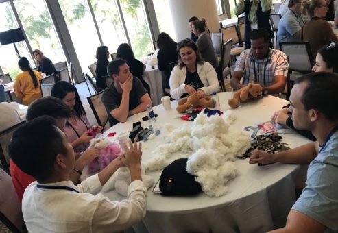 A group of individuals sits around a table working on a Team Teddy Rescue Bear event. Participants are stuffing teddy bears and organizing materials like cotton filling, bear parts, and accessories. The room is filled with other groups, all engaged in similar activities.