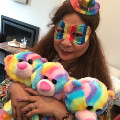 A woman dressed festively in a rainbow-colored mask and a mini hat, holding three brightly colored rainbow teddy bears. She has a warm expression as she hugs the bears. The setting appears to be a fun and playful environment, part of a charitable team building event. thumbnail